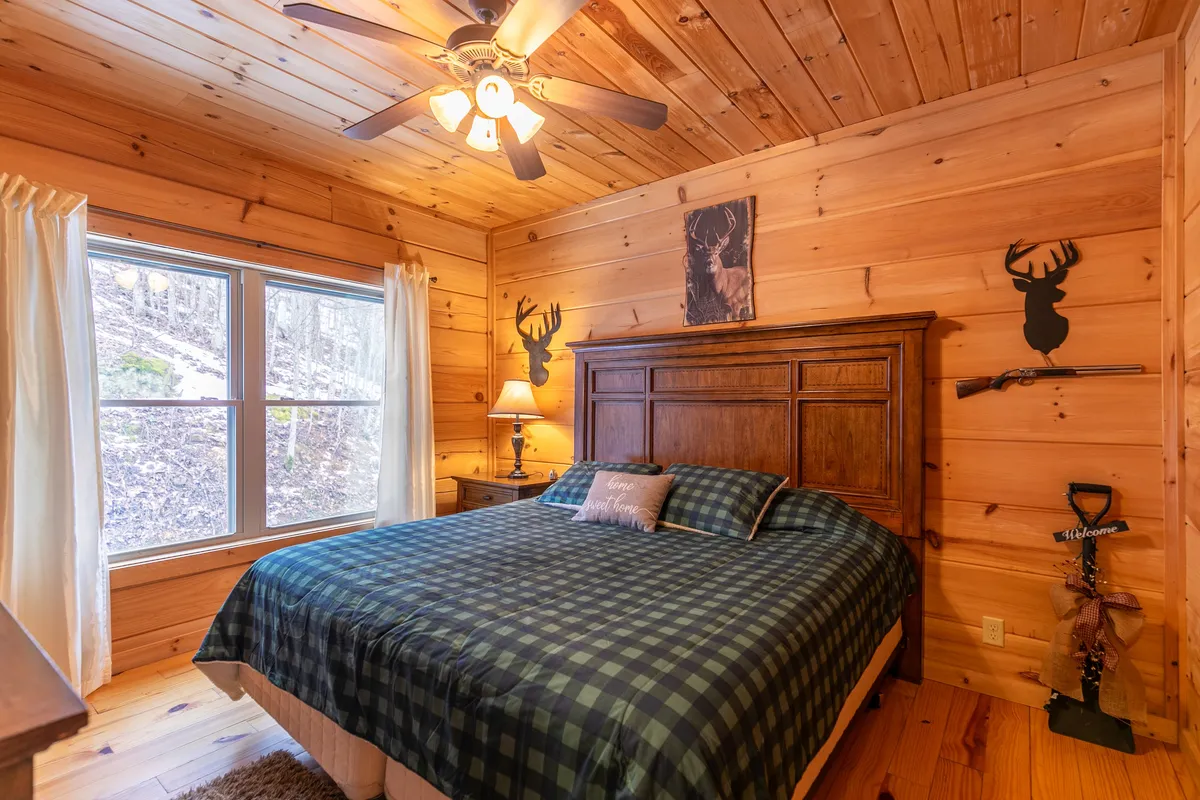 A cozy bedroom featuring a bed with plaid bedding, a wooden headboard, decorative antler wall art above the bed, and a window letting in natural light. The rustic decor and warm textures evoke comfort and blend with the cabin’s mountain charm.