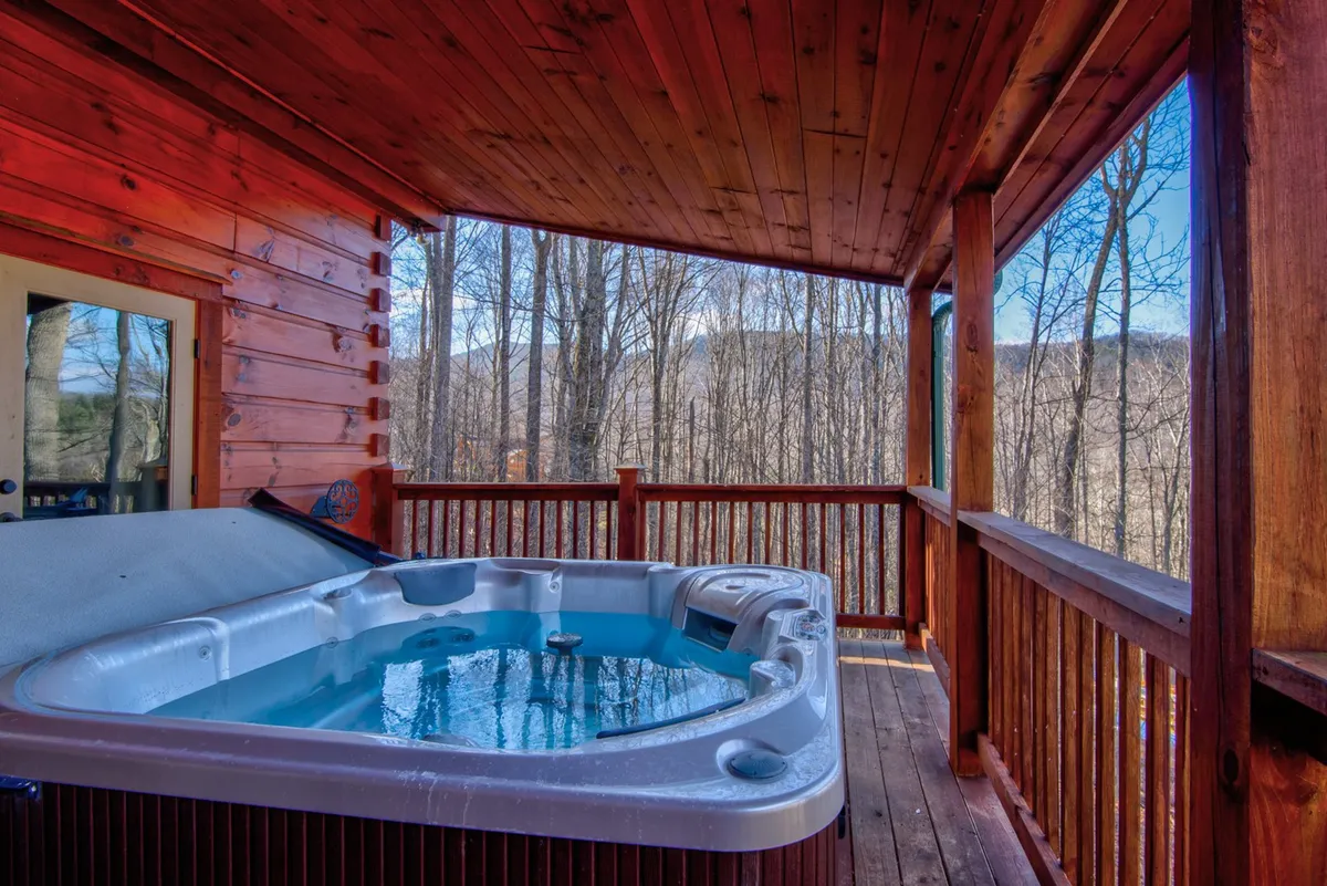 An inviting outdoor hot tub sits on a wooden deck, surrounded by lush trees with a scenic mountain view visible on the horizon. The space is tranquil, offering a private oasis for relaxation and socializing under the open sky.