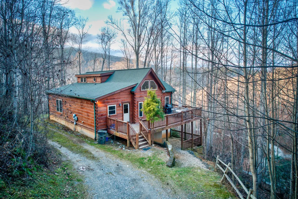 This exterior shot highlights the wooden cabin nestled among trees, with a visible deck and a natural pathway leading towards the entrance. The surroundings evoke peaceful nature walks, sunrises, and evenings spent together outdoors.