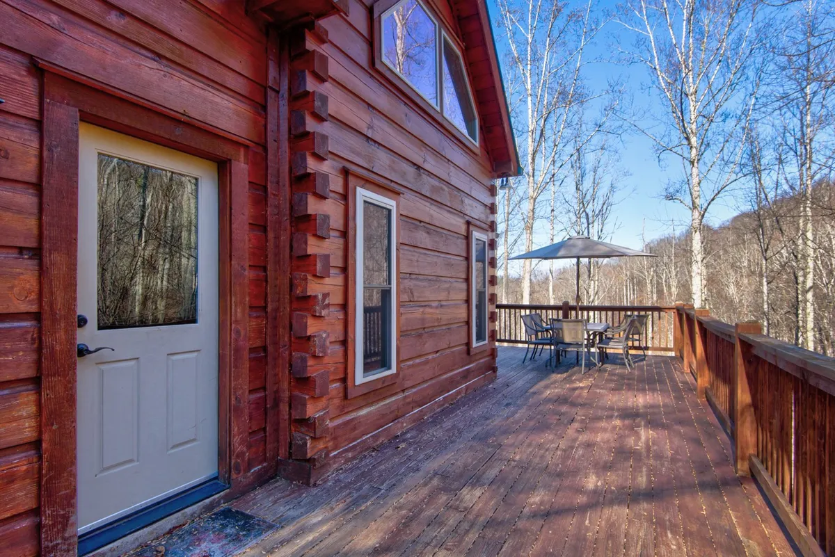 A wooden deck with comfortable outdoor seating, large windows looking into the cabin, and trees all around. The tranquil setting is ideal for enjoying coffee, al fresco meals, or simply soaking in the peaceful mountain atmosphere.