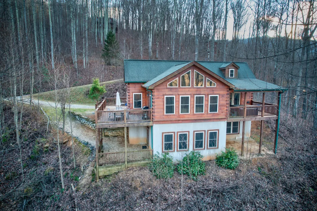 The image shows the wooden exterior of the house, featuring large windows and a spacious deck surrounded by trees. The deck is set up for outdoor gatherings and barbecues, perfectly positioned to catch sunset views and starry nights in the clear mountain sky.