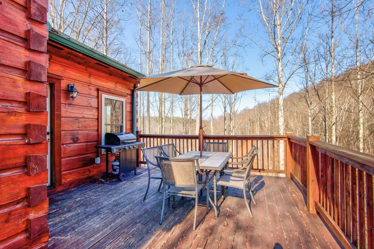 A wider exterior view showcasing a wooden deck fitted with a grill, dining table with chairs, and a pergola umbrella. The scene is perfect for barbecuing and socializing, with the surrounding woods creating a private, relaxing environment.