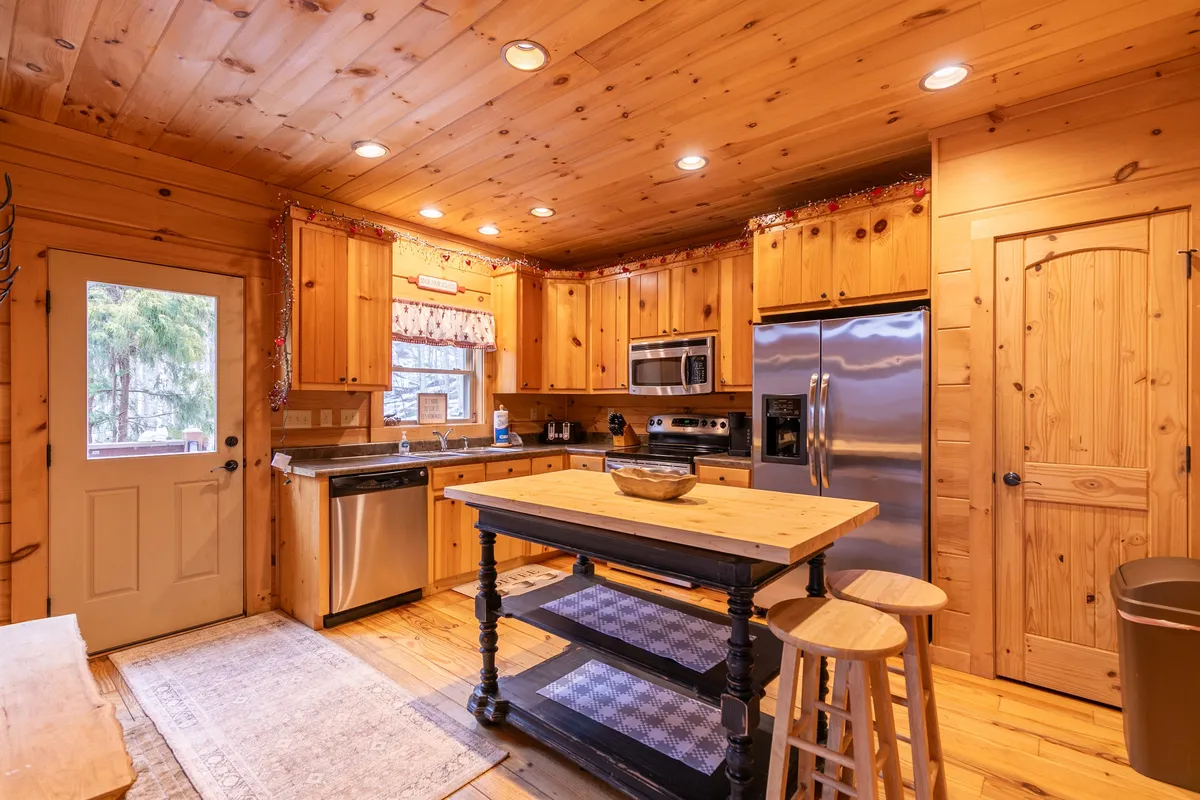 The image shows a warmly lit kitchen with a prominent wooden island, stainless steel appliances, granite countertops, and ample cabinetry. A large window allows natural light to stream in, highlighting the welcoming and functional design perfect for group meals and gatherings.