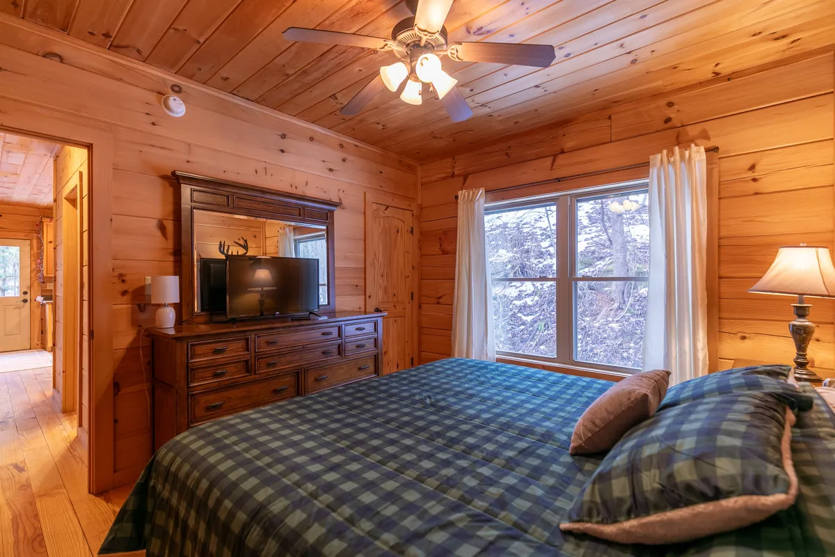 A spacious bedroom with a bed covered in a checkered comforter, a large window providing ample natural light, a wooden dresser topped with a TV, and a ceiling fan overhead. The room feels comfortable, inviting, and ideal for relaxation or quiet time.