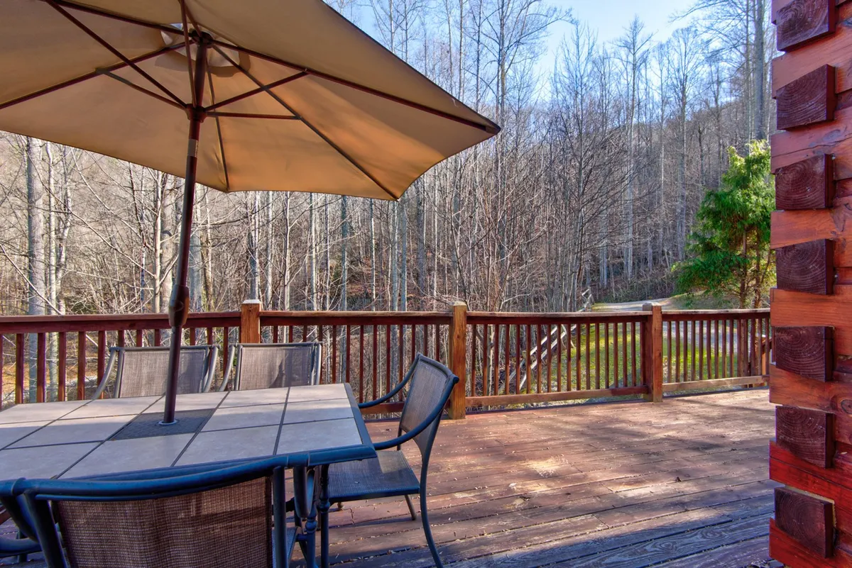 An inviting view of an outdoor dining table with an umbrella set on a wooden deck, surrounded by trees. The table is ready for al fresco meals, with ample seating for guests and a peaceful backdrop of nature.