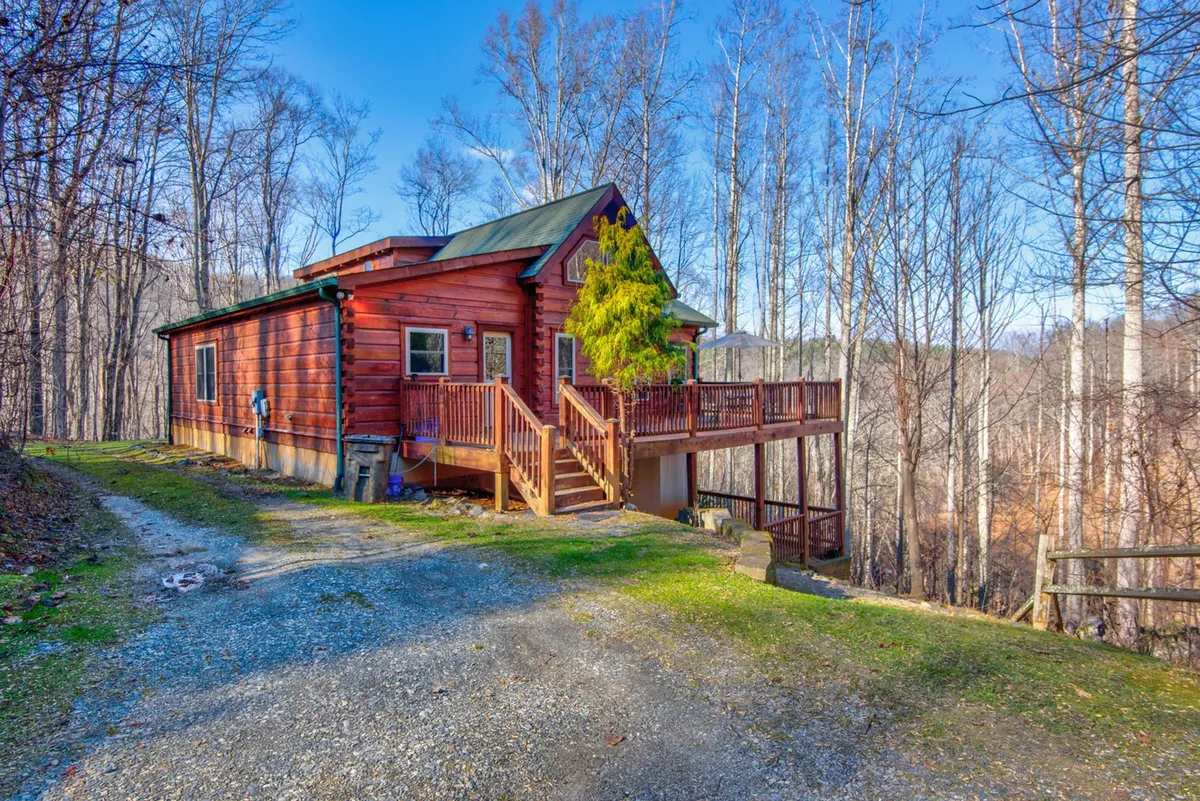 A charming log cabin nestled among mature trees, featuring a spacious wooden deck with seating. The cabin is surrounded by nature, with a gravel pathway leading up to the entrance, offering a sense of privacy and tranquility in a picturesque wooded setting.
