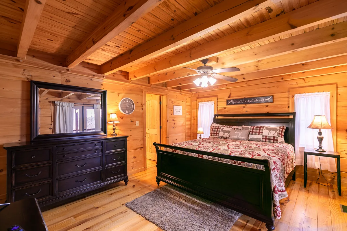 A warmly lit bedroom with a patterned comforter on the bed, a dark wooden dresser, lamps positioned on either side for soft lighting, and a large mirror reflecting the room’s inviting ambiance. The overall feeling is of coziness and privacy.