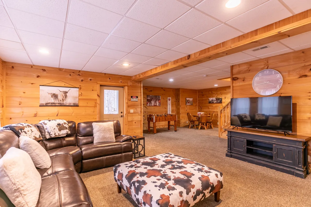 A spacious entertainment area with a leather sectional sofa, cowhide ottoman, large television, and adjacent wooden dining table. The arrangement invites movie marathons, socializing, and shared activities in a warm, rustic setting.
