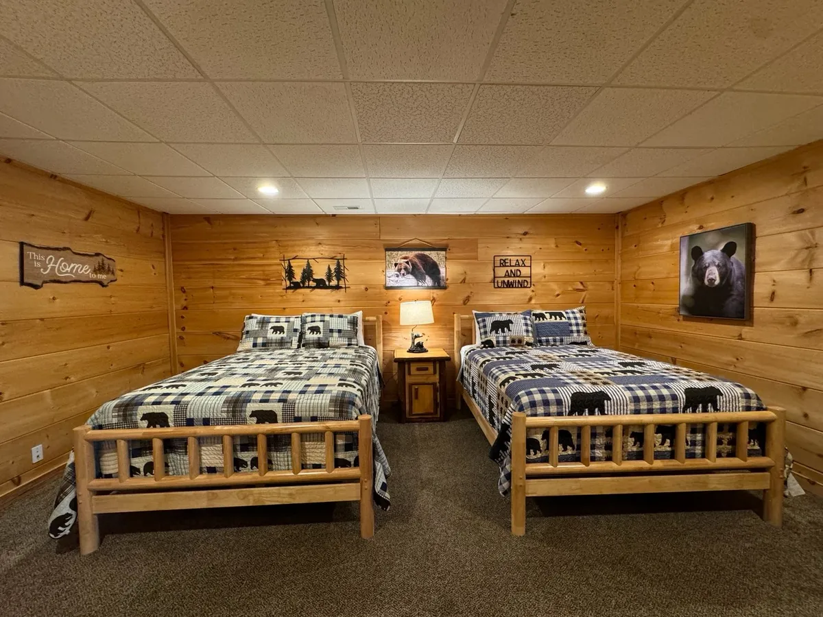 A bedroom with two wooden beds dressed in plaid blankets, nature-themed wall art depicting bears and forest scenes, and a wooden side table with a lamp between the beds. The decor emphasizes warmth, family togetherness, and a connection to nature.